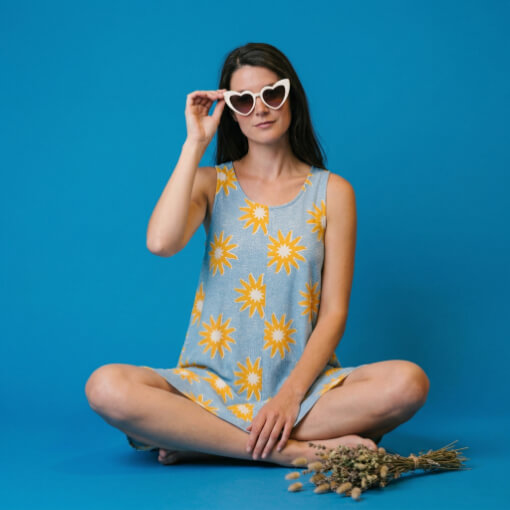 Robe Rava Esprit de la Mer. Une femme vêtue d'une robe bleue à motifs floraux ajuste ses lunettes de soleil blanches en forme de cœur, assise les jambes croisées.